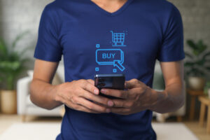Pessoa usando smartphone com ícone de compra online “BUY” e carrinho de compras projetados sobre a camiseta azul.